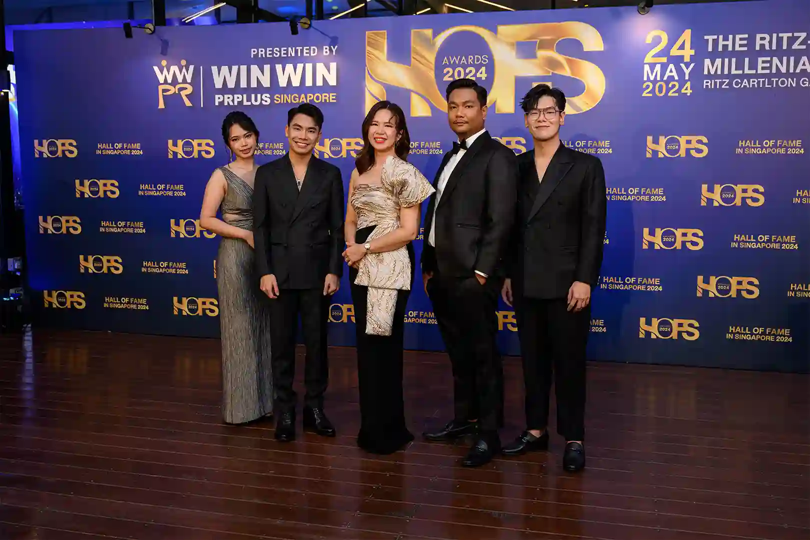 ภาพกลุ่มผู้ชนะรางวัล HOF Awards 2024 ที่งานประกาศรางวัล ณ โรงแรม Ritz Carlton, Singapore เมื่อวันที่ 24 พฤษภาคม 2024 บุคคลในภาพแต่งกายด้วยชุดทางการ หญิงหนึ่งในชุดเดรสสีเงิน ชายสองคนในชุดสูทสีดำ และหญิงอีกคนในชุดเดรสสีทอง-เงิน พื้นหลังเป็นแบนเนอร์สีน้ำเงินที่มีข้อความแสดงถึงงาน Hall of Fame Awards 2024 ที่จัดขึ้นโดย WIN WIN PRPLUS Singapore ภาพนี้เน้นถึงความสำเร็จและการยอมรับในระดับนานาชาติของผู้ชนะรางวัล